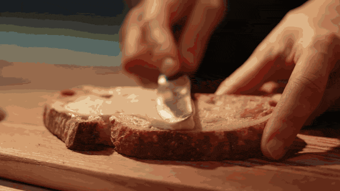 Butter is spread on toasted bread, then topped with tomatoes and basil, in warm golden light