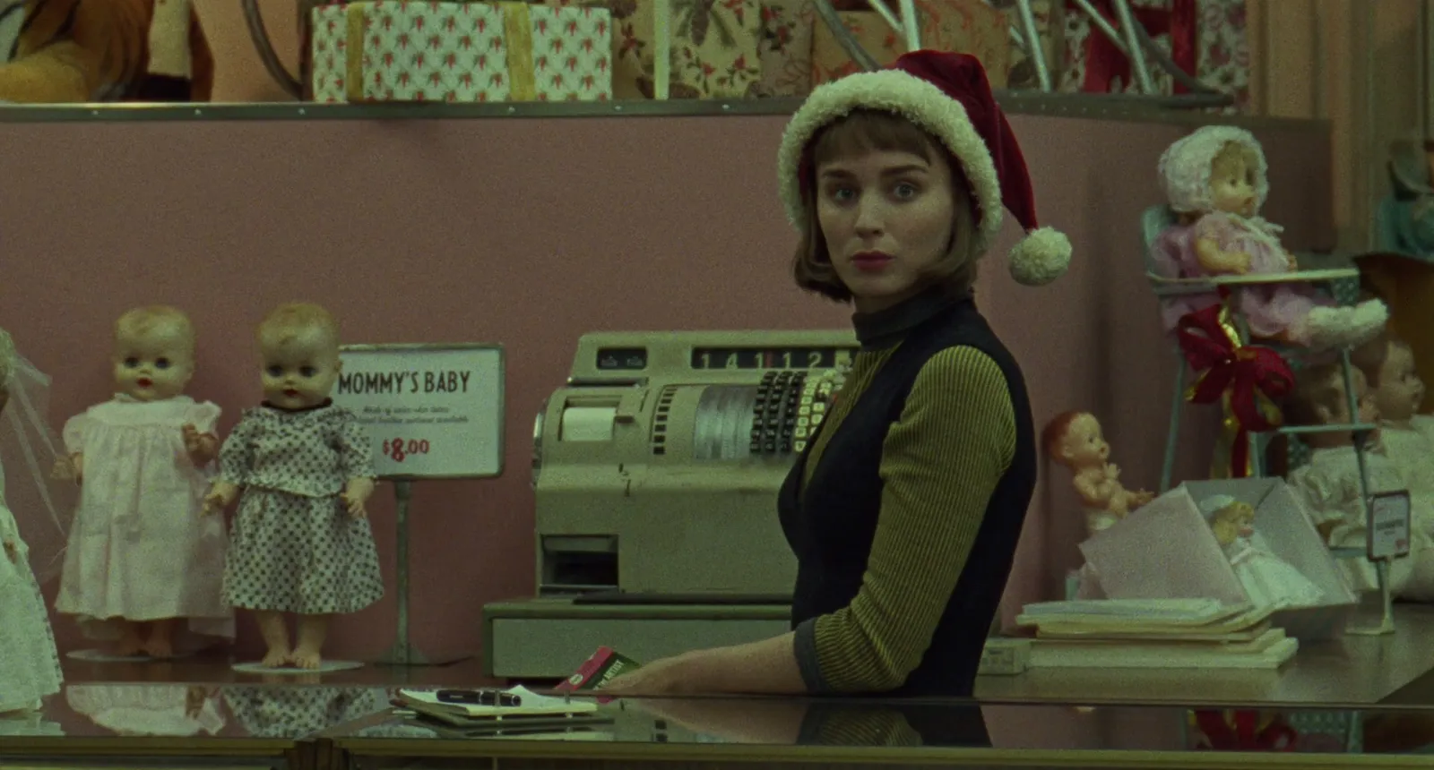 A shopgirl in a Santa hat behind a department-store toy counter in dusty pink, olive and cream