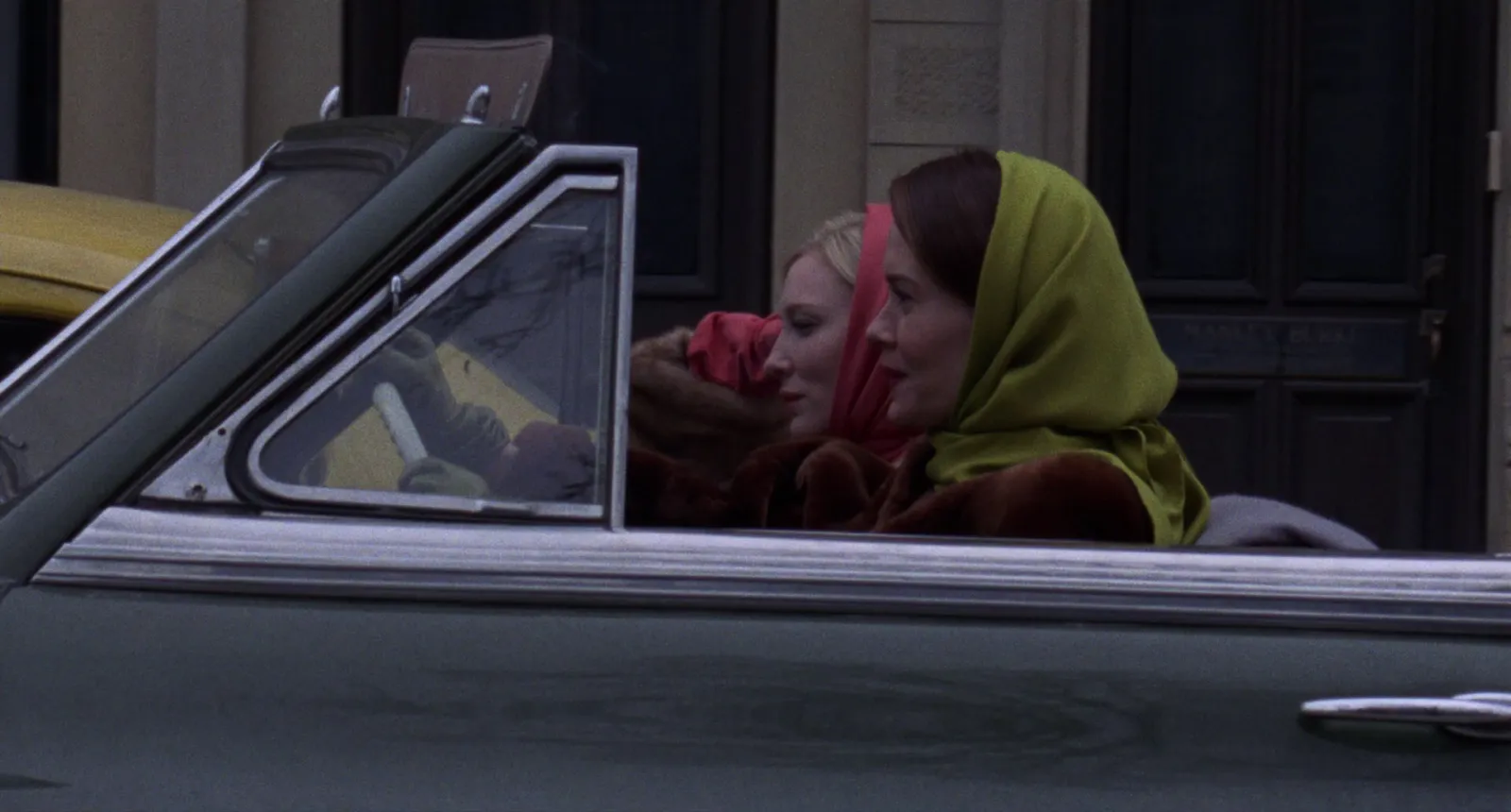 Two women in a grey convertible, one in a mustard headscarf and one in red, muted greens and greys