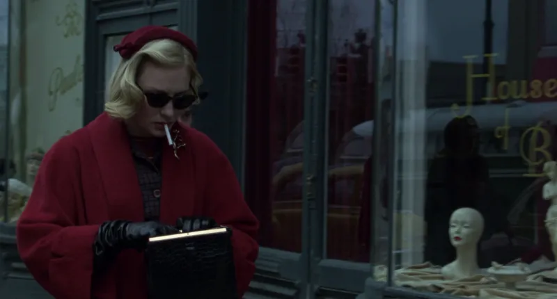 A blonde woman in a red coat, red beret and dark glasses outside a shop window, red against slate grey, from Carol