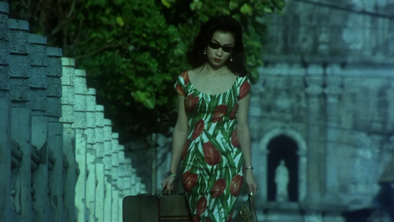 A woman in sunglasses and a red-and-green tulip print dress walking past stone balustrades in teal shade