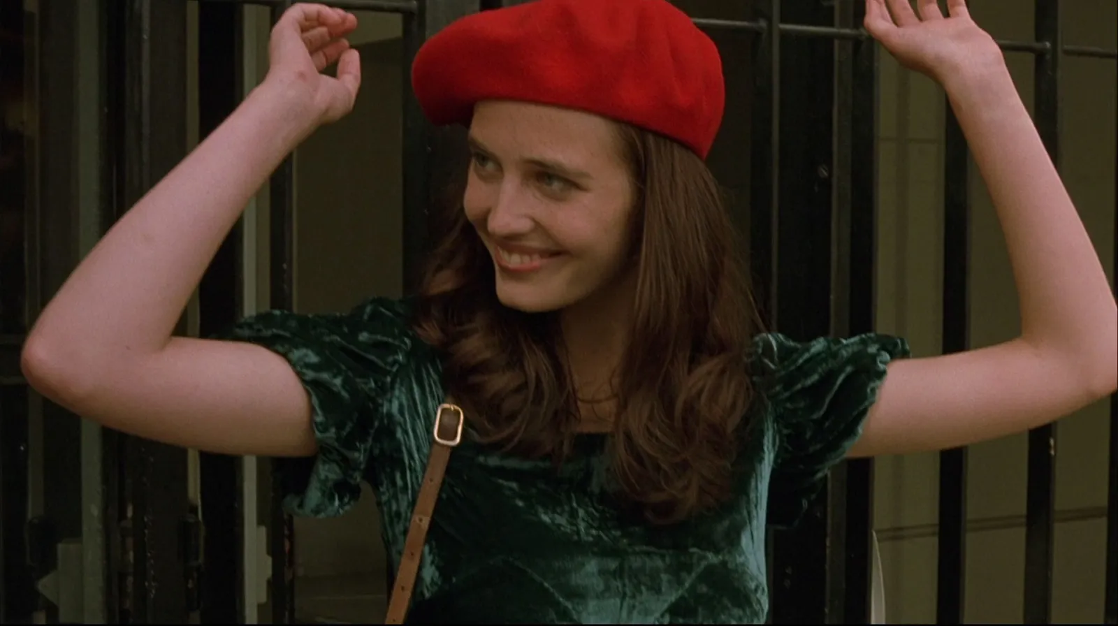 A young woman in a red beret and green velvet dress, arms raised against iron gates