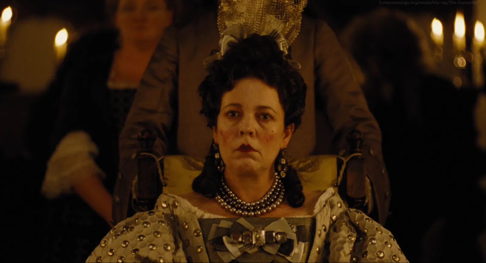 A queen enthroned in a gold studded gown with candles behind her