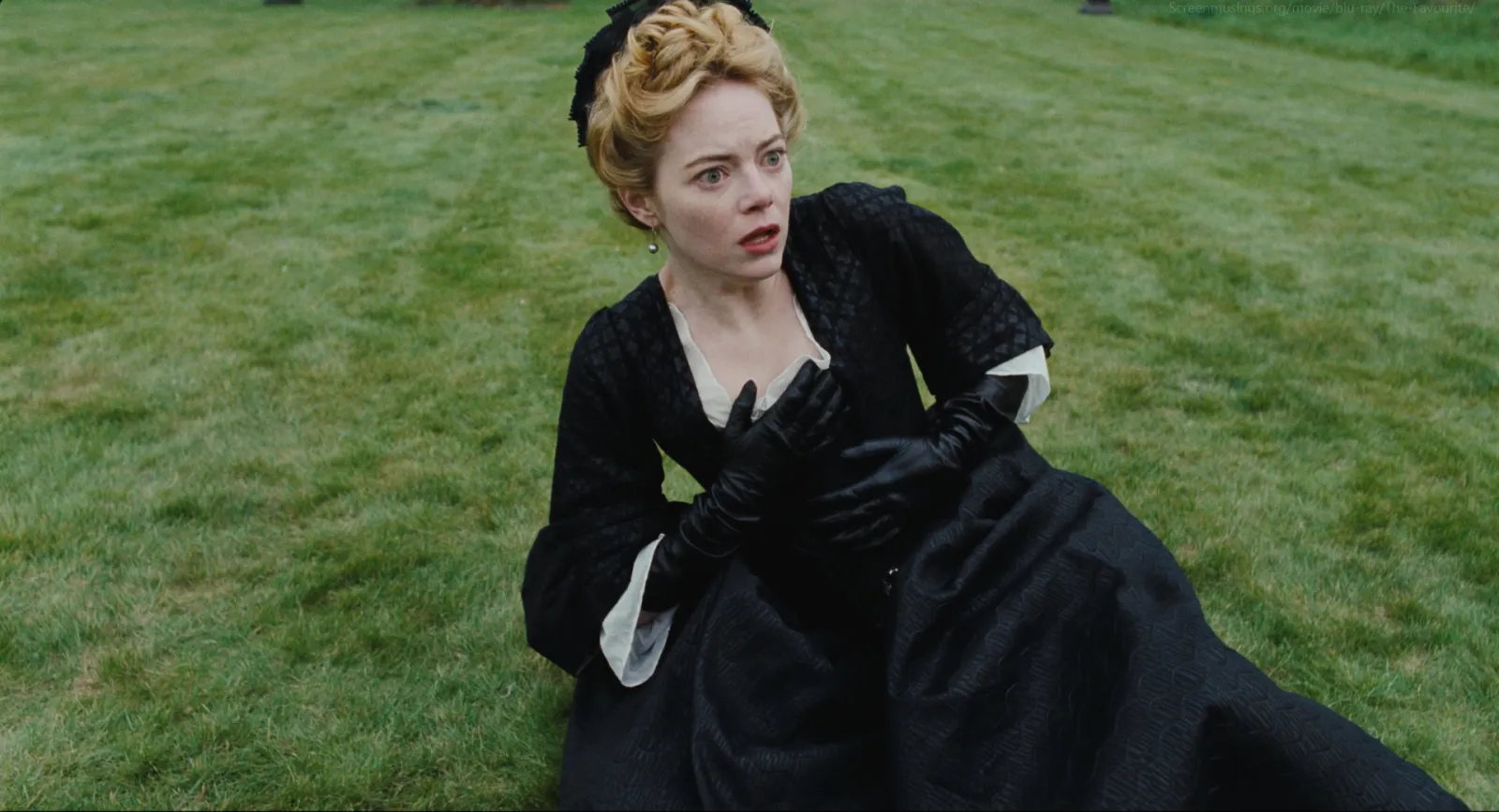 A woman in a black gown and black gloves sprawled on a flat green lawn under overcast light