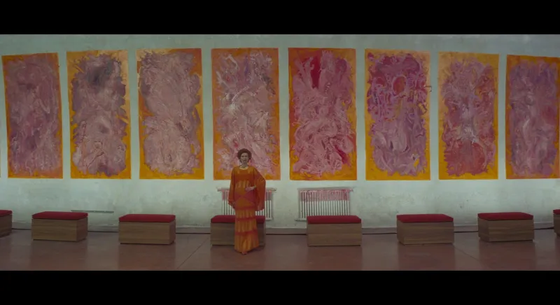 A wall of pink canvases with a figure in an orange robe standing dead centre, perfectly symmetrical