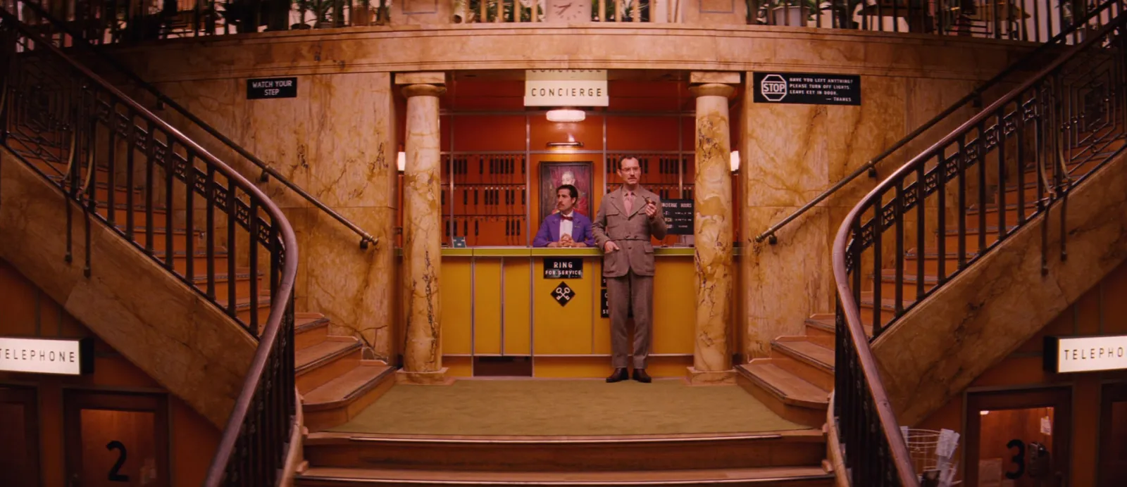 A grand hotel lobby with twin staircases and one tiny figure at the desk