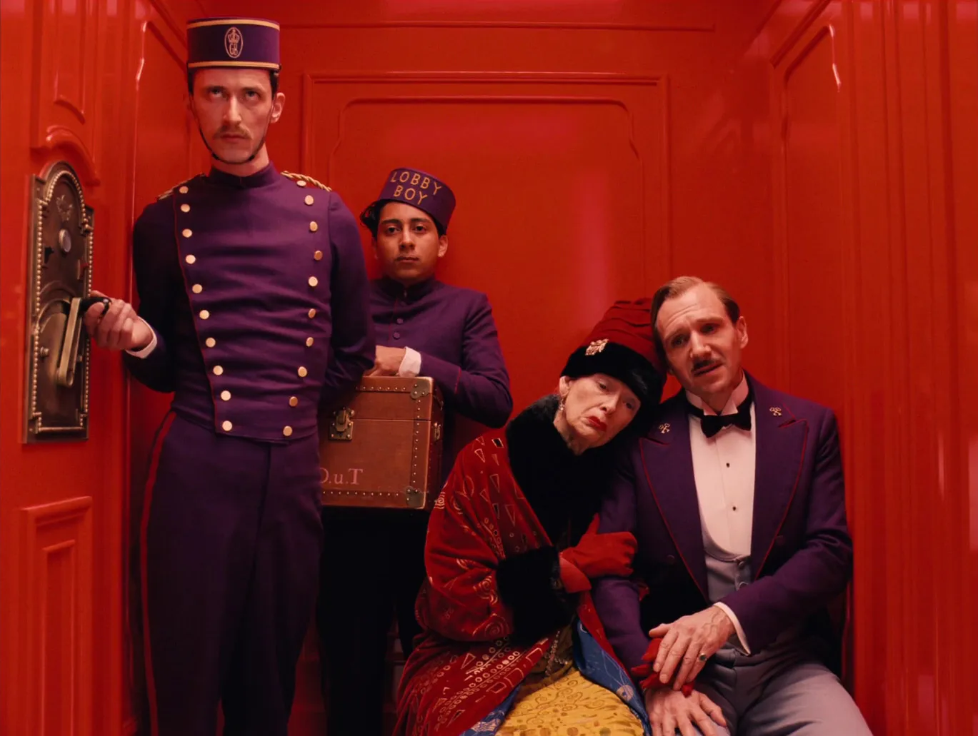 A crowded hotel lift lit deep red, staff in purple uniforms