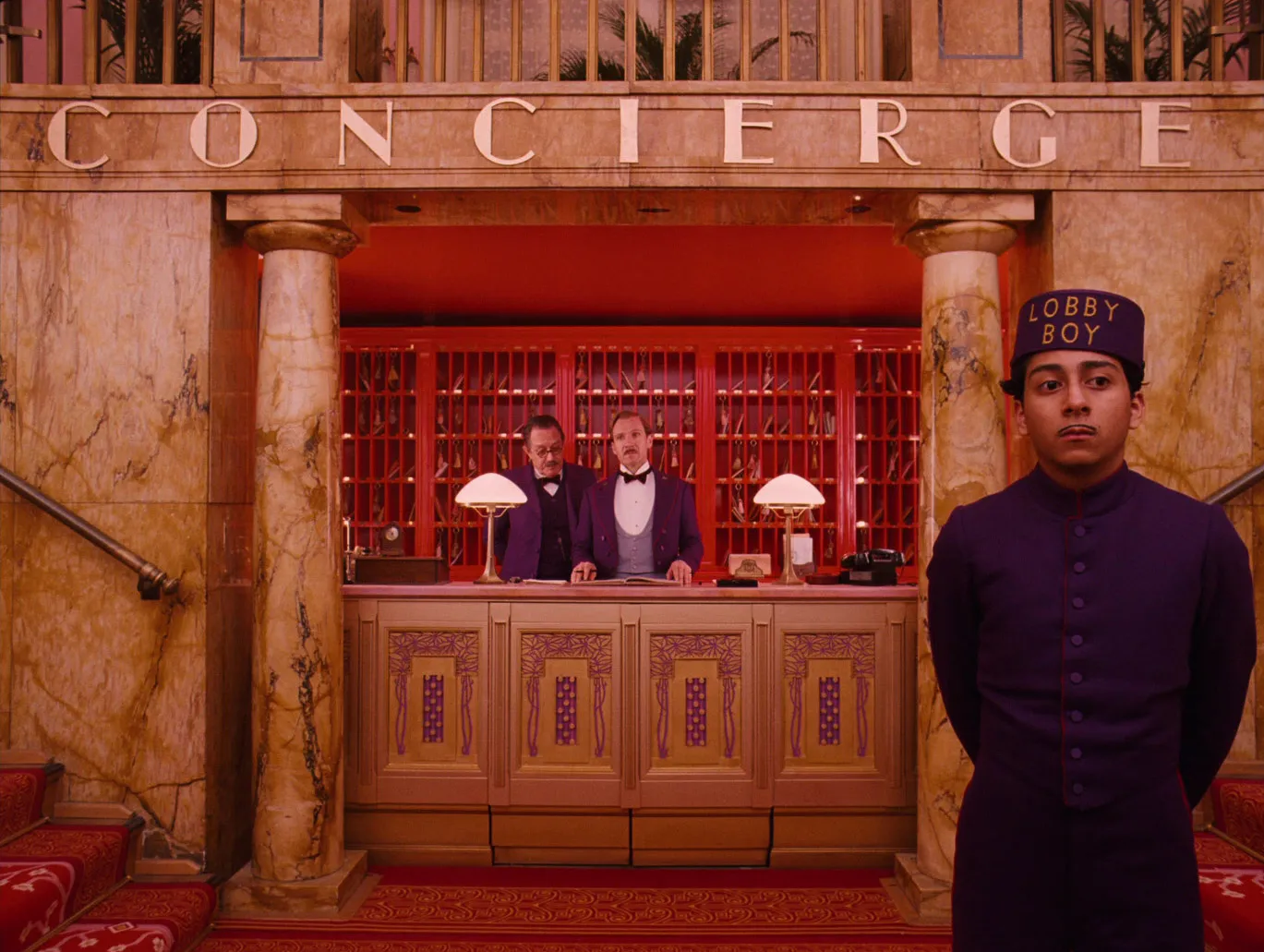 A hotel concierge desk in red and gold under a CONCIERGE sign, with uniformed staff lined up symmetrically behind it