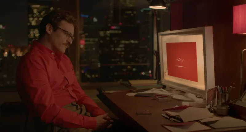 A man in a red shirt sitting at a desk, his screen glowing in a warm red and pink room