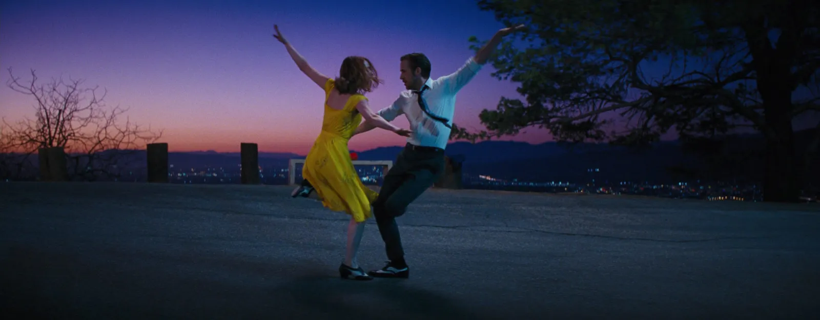 A couple dancing on a hillside at purple-blue dusk, the city lights spread out below them