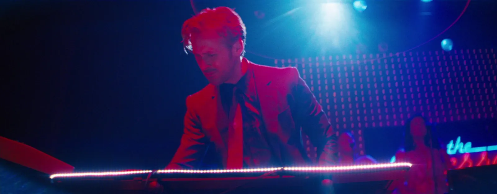 A pianist in a red jacket under red and blue light, a neon sign behind