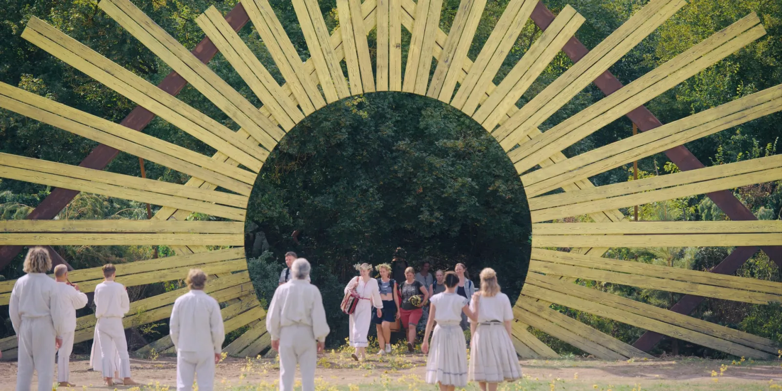 A group dressed in white walks through a huge wooden sunburst arch into a forest, in pale yellow and green, from Midsommar