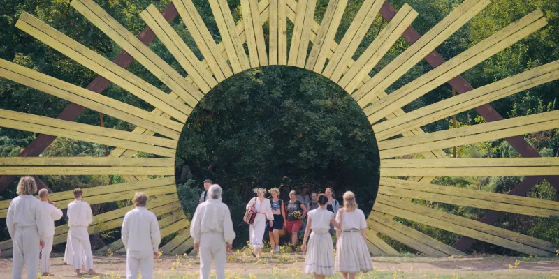A group dressed in white walks through a huge wooden sunburst arch into a forest, in pale yellow and green, from Midsommar