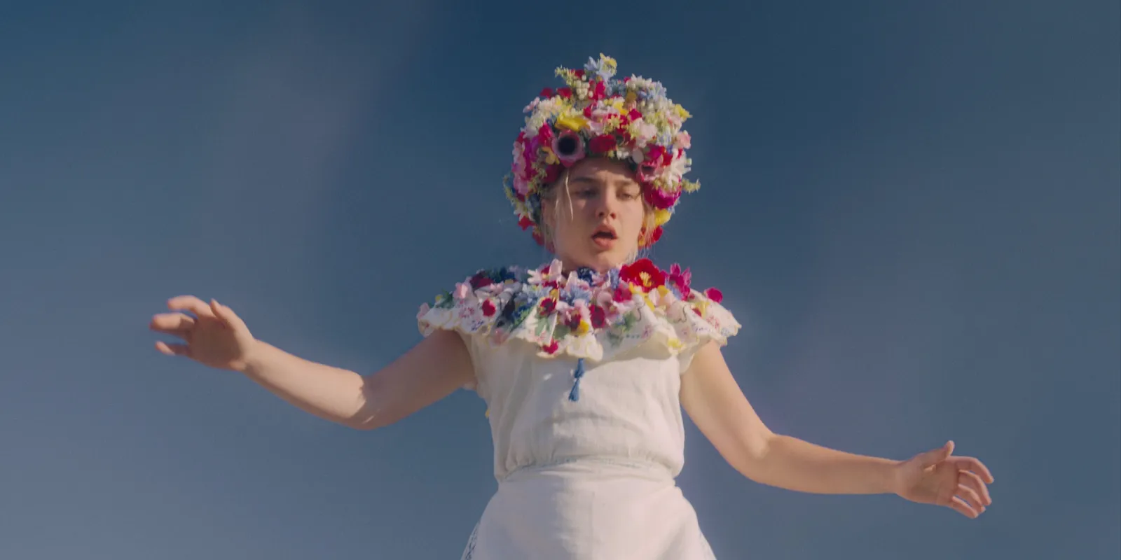 A girl in a white dress and flower crown with her arms out, shot from below against a clear blue sky