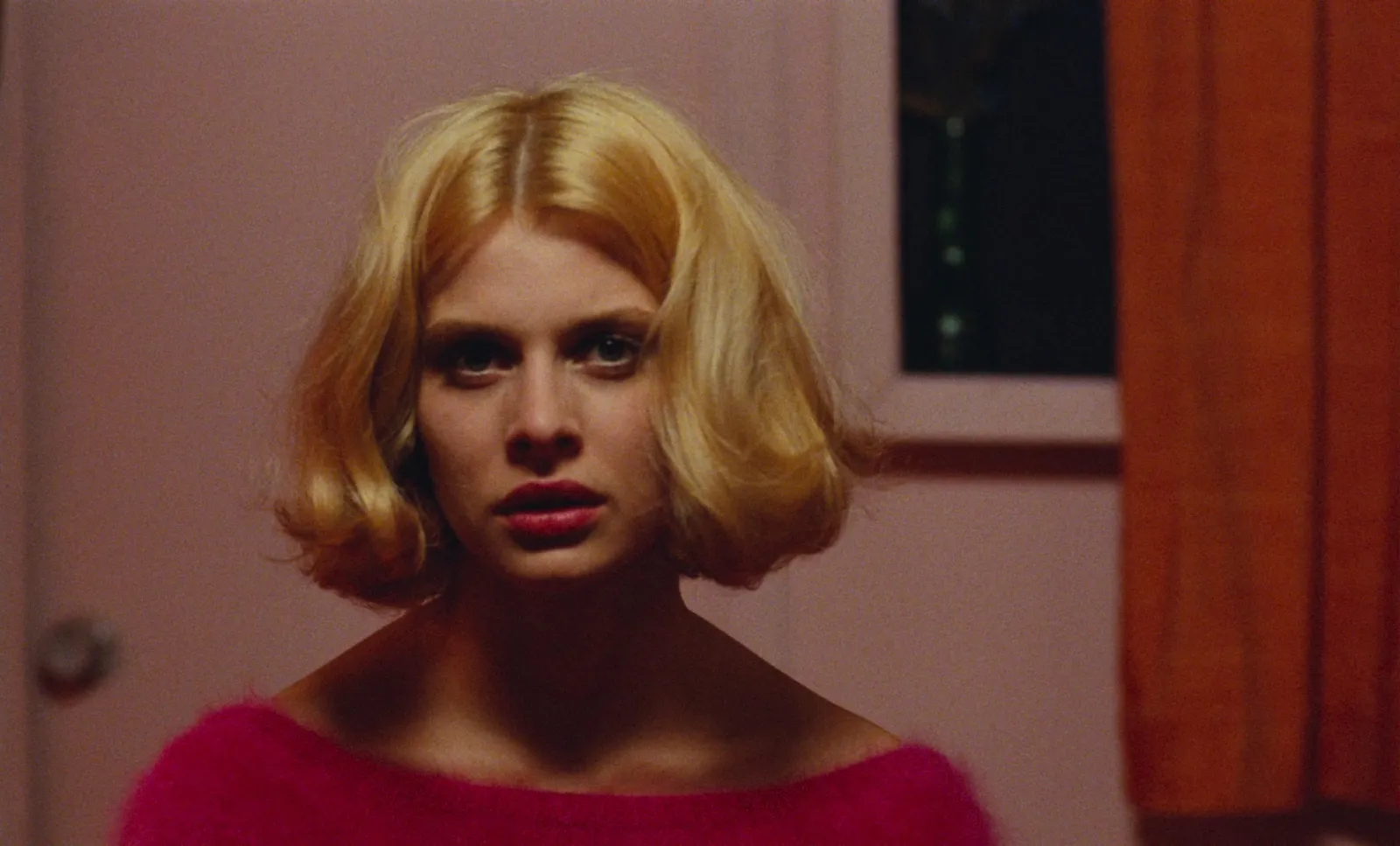 A woman with a blonde bob in a pink mohair sweater against a peach wall and orange curtain