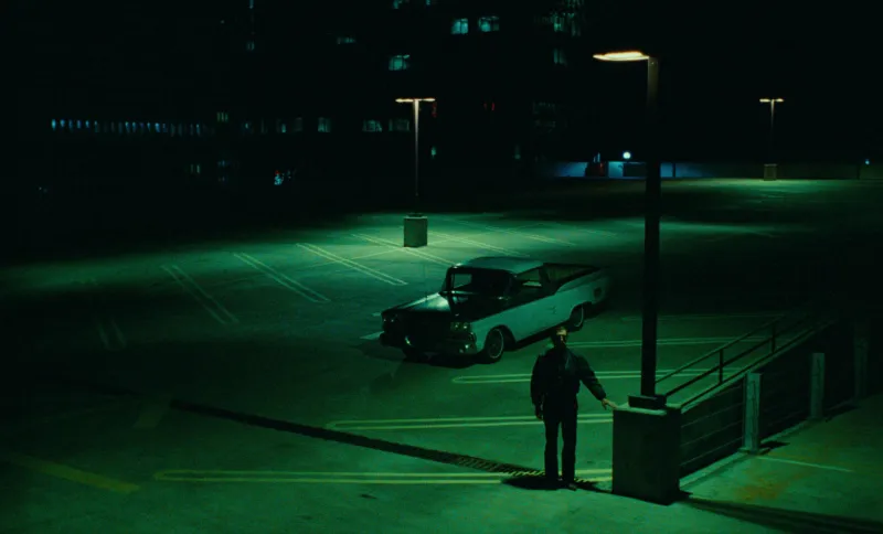 A man beside an old car in an empty car park at night, lit sodium green, from Paris, Texas