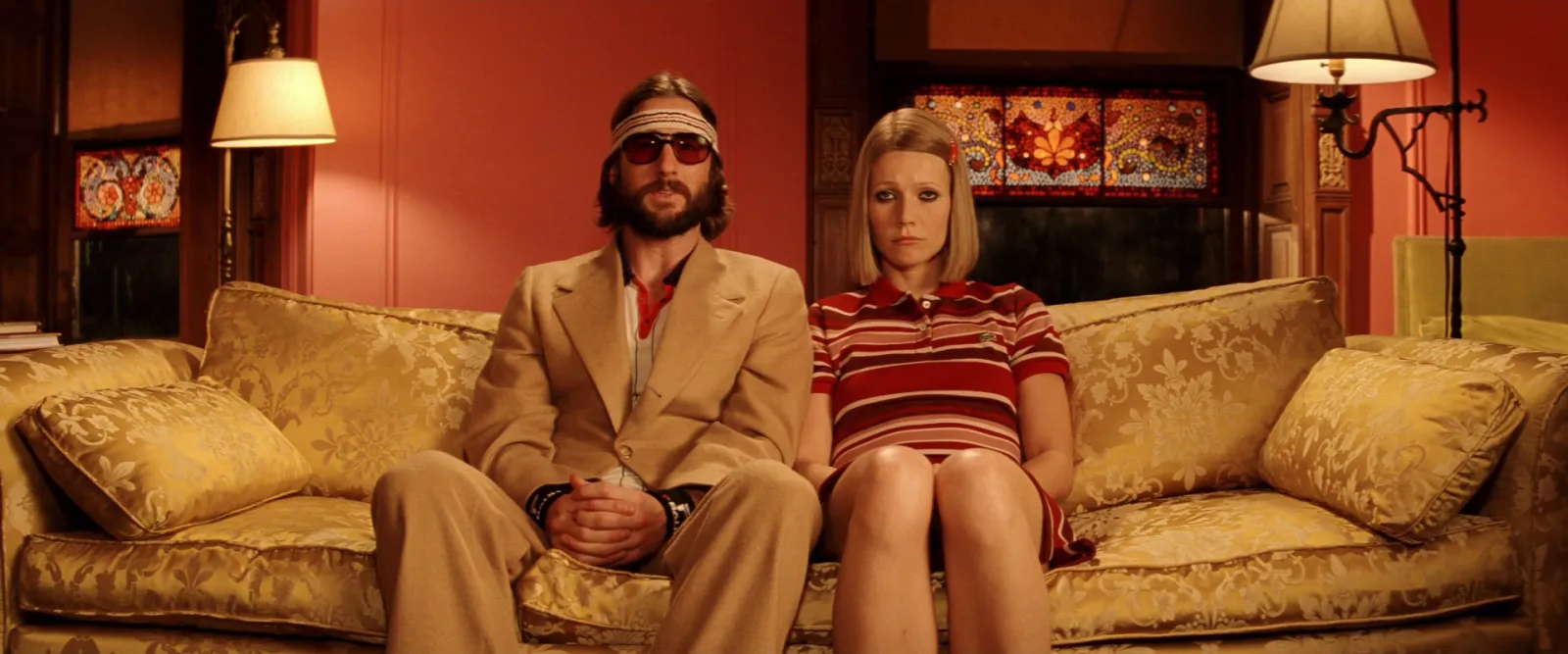 A couple sitting apart on a gold sofa in a red-brown room, framed symmetrically