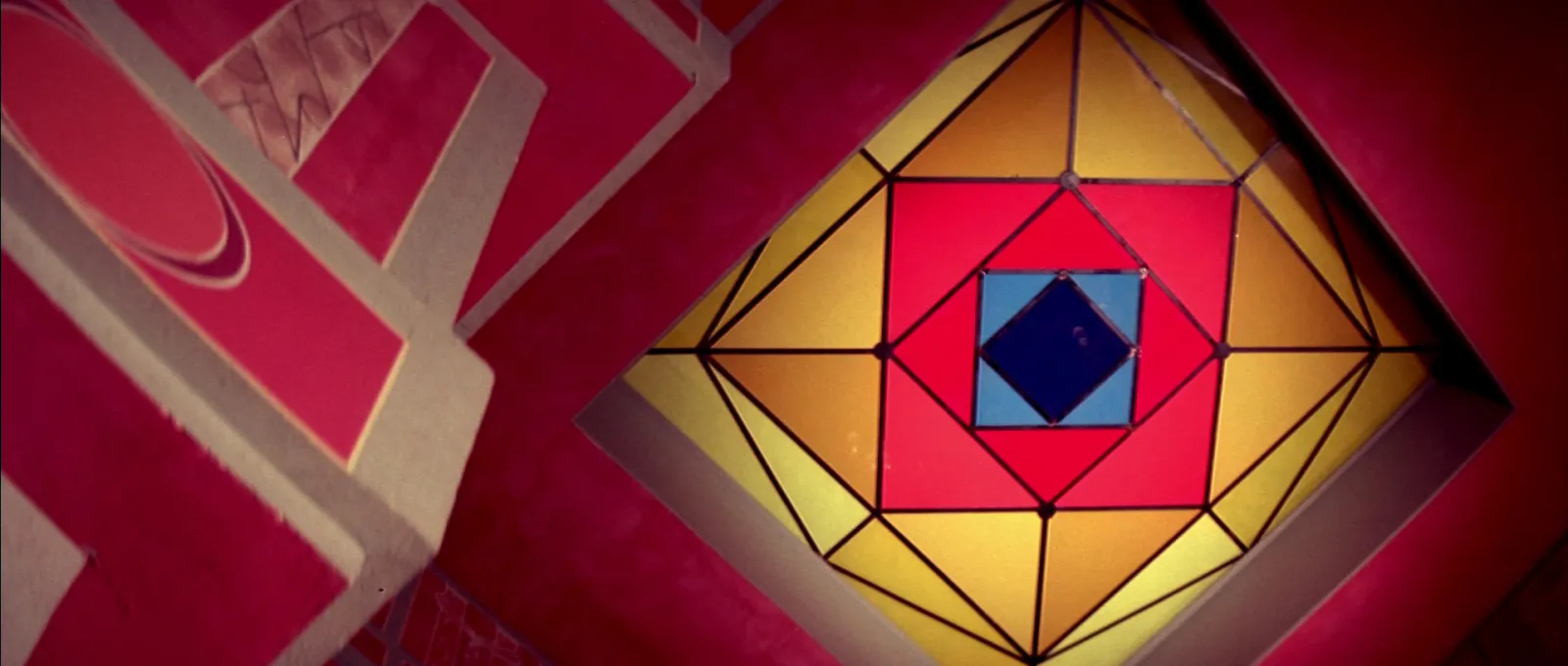 A geometric stained glass ceiling panel in yellow, red and blue set into a red wall