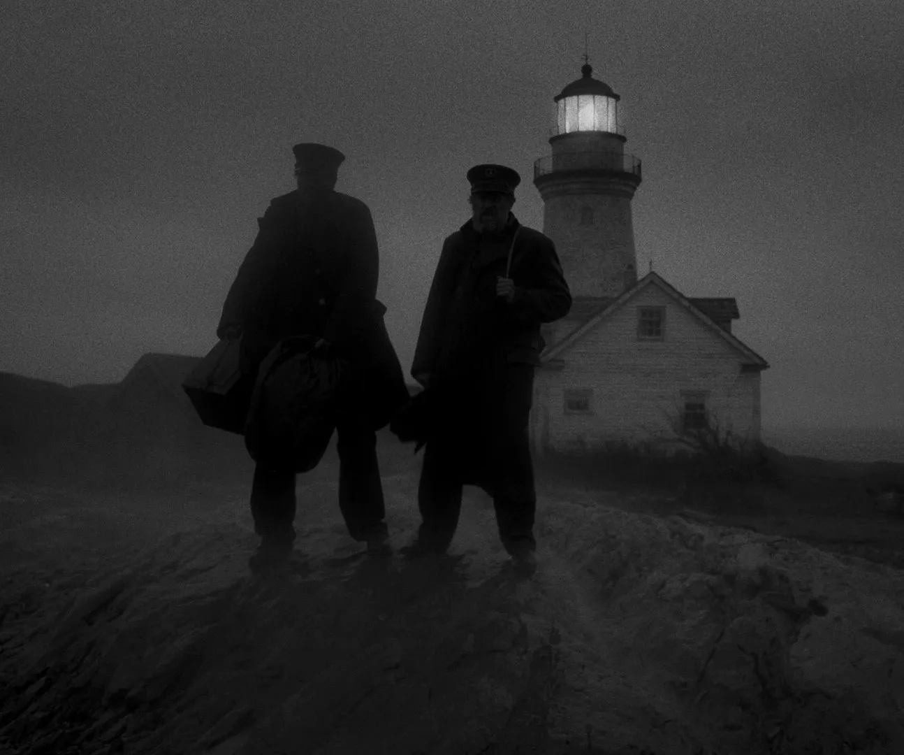 Two lighthouse keepers standing in fog in front of the lighthouse, in black and white